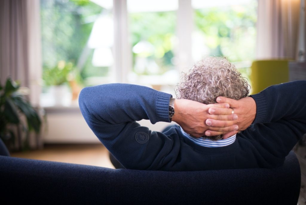 Mann sitzt mit hinter dem Kopf verschränkten Armen auf dem Sofa